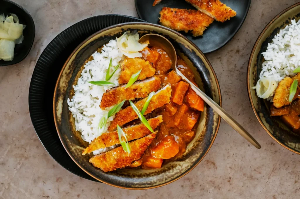 A bowl of Japanese curry with white rice and crispy breaded chicken cutlets, garnished with sliced green onions and pickled ginger on the side.