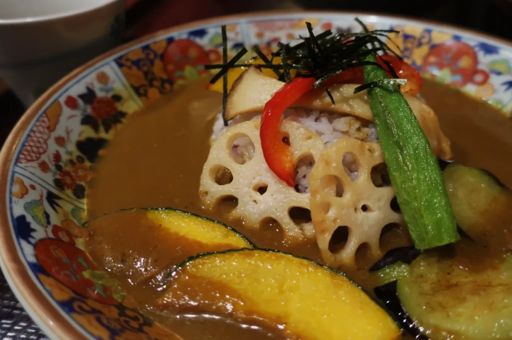 A colorful bowl of Japanese curry features rice topped with sliced lotus root, red pepper, okra, seaweed, and mushrooms, on a vibrant patterned plate.