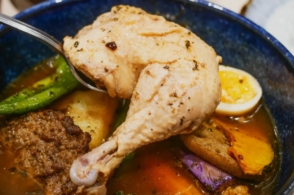 A flavorful blue bowl of curry soup with a tender chicken leg, boiled egg, vibrant vegetables, and a savory patty, creating a warm, hearty scene.