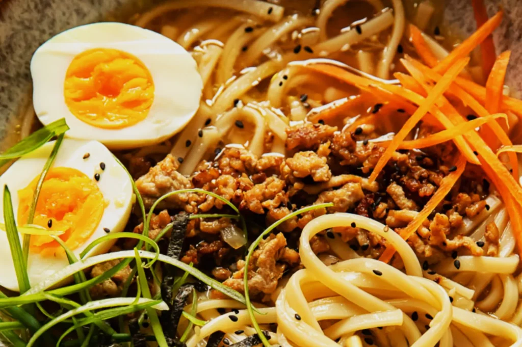 Bowl of ramen with sliced boiled eggs, minced pork, and noodles, garnished with green onions, carrot strips, and black sesame seeds.