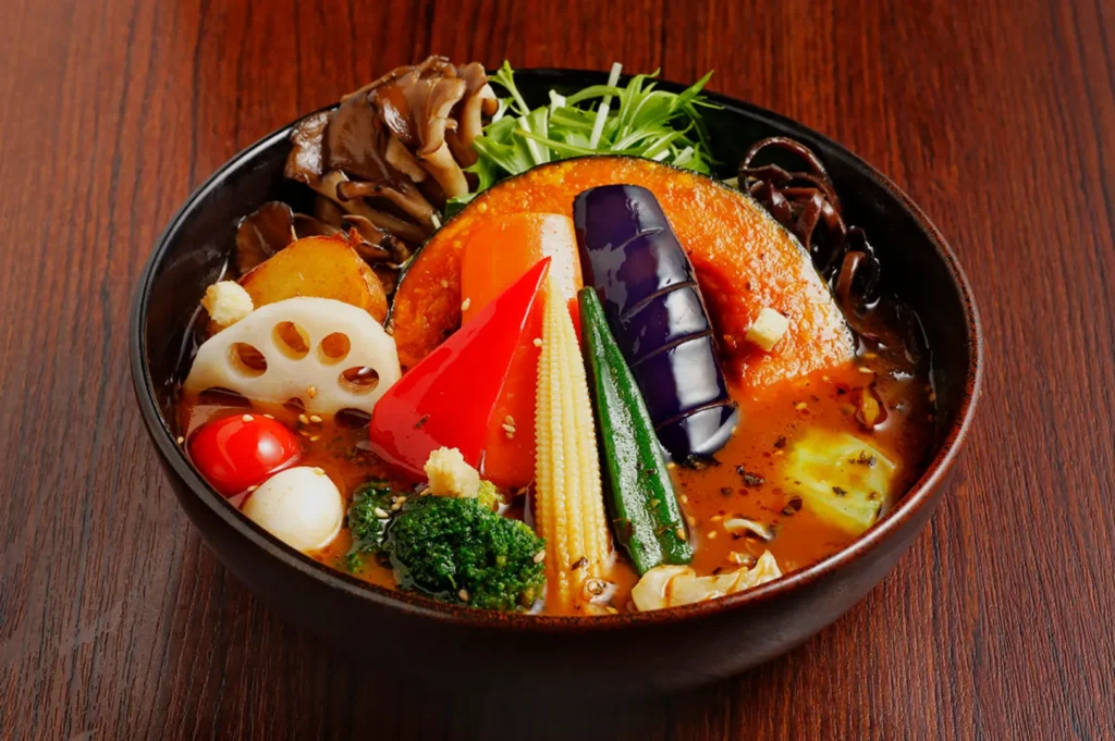 A bowl of vibrant vegetable curry on a wooden table. Includes pumpkin, eggplant, bell pepper, lotus root, baby corn, mushrooms, and greens in rich broth.