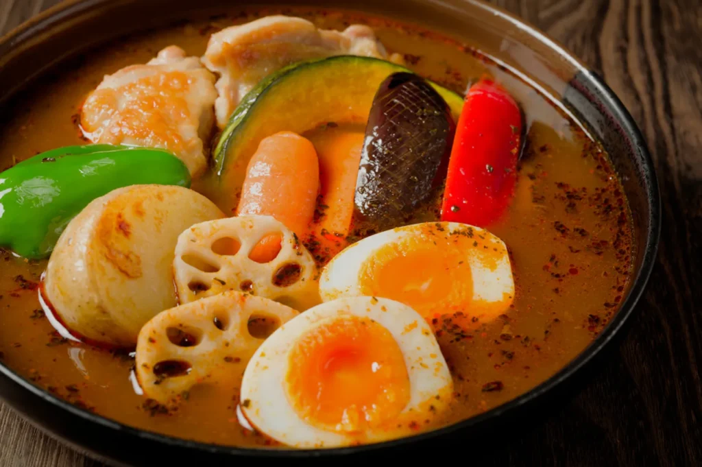 A colorful bowl of Japanese curry soup with sliced boiled eggs, lotus root, green pepper, carrot, pumpkin, and chicken in rich broth on a wooden table.