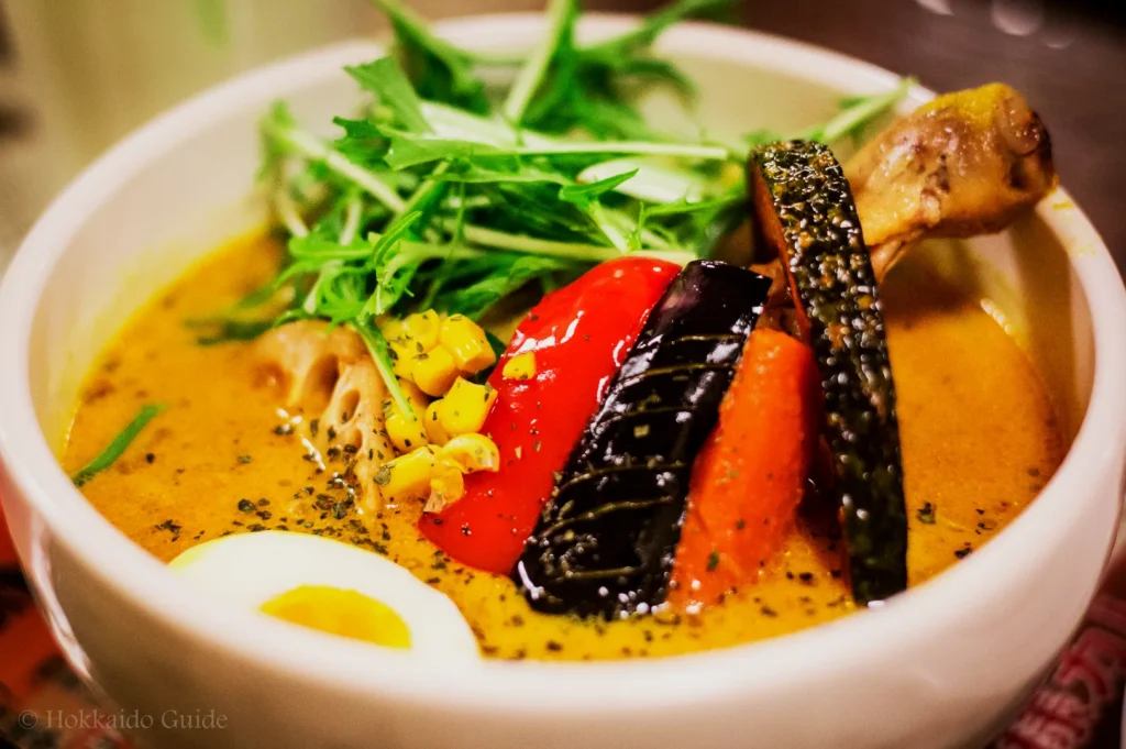 A vibrant bowl of soup curry with colorful vegetables like corn, red pepper, eggplant, and carrots, accompanied by a boiled egg and fresh greens.