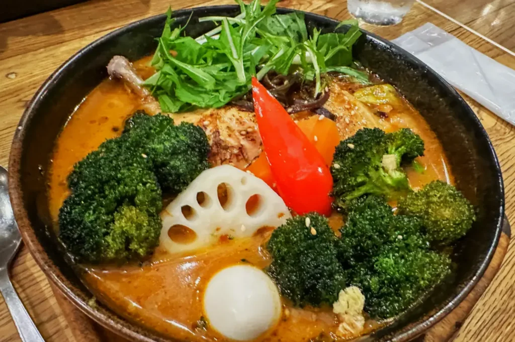 A steaming bowl of vibrant curry soup with broccoli, lotus root, a boiled egg, a red pepper, greens, and savory broth on a wooden table.
