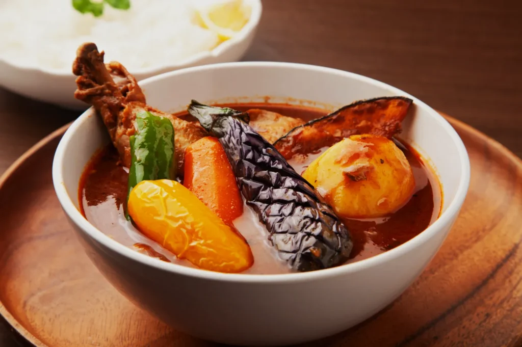 A bowl of rich, spicy curry features colorful vegetables like eggplant, bell peppers, and a potato with a chicken drumstick, sitting on a wooden tray. In the background, a bowl of steamed white rice garnished with a sprig of green herb. The scene emits a warm, inviting vibe.