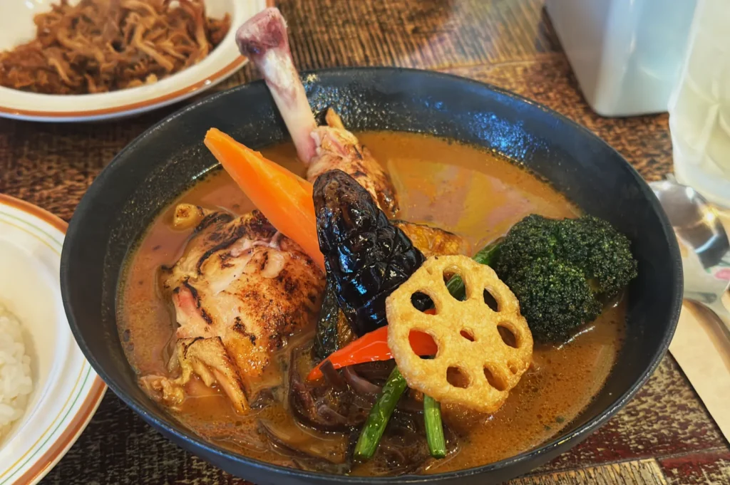 A vibrant bowl of curry with chicken leg, carrots, eggplant, broccoli, and lotus root in rich broth, set on a wooden table, exuding warmth.