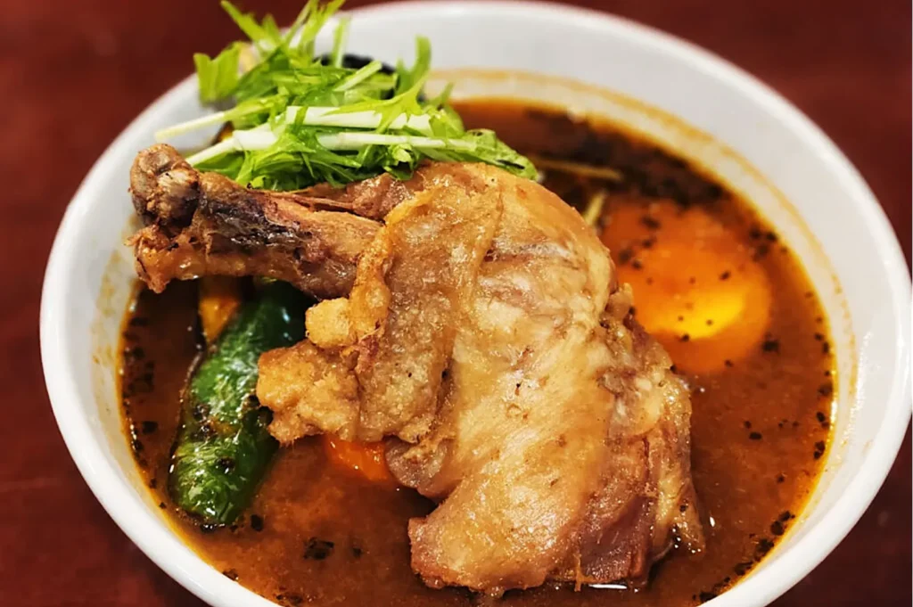 A bowl of rich, brown curry soup features a crispy, golden chicken leg, green peppers, and carrots, garnished with fresh green herbs on top.