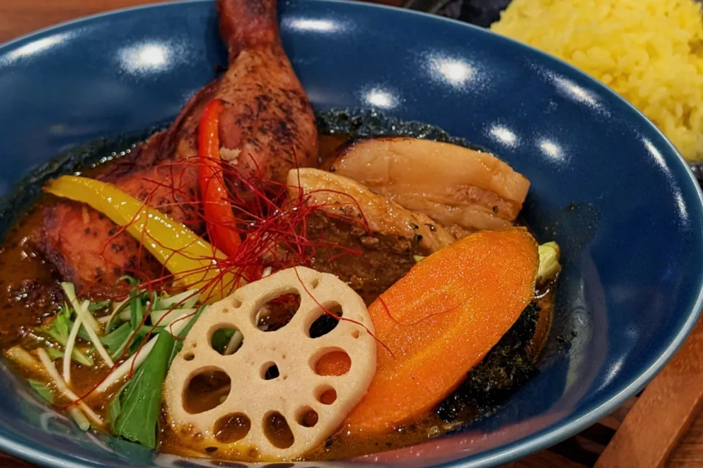 A vibrant bowl of Japanese curry with a grilled chicken drumstick, carrot slices, lotus root, yellow and red peppers, and greens. Served with yellow rice.