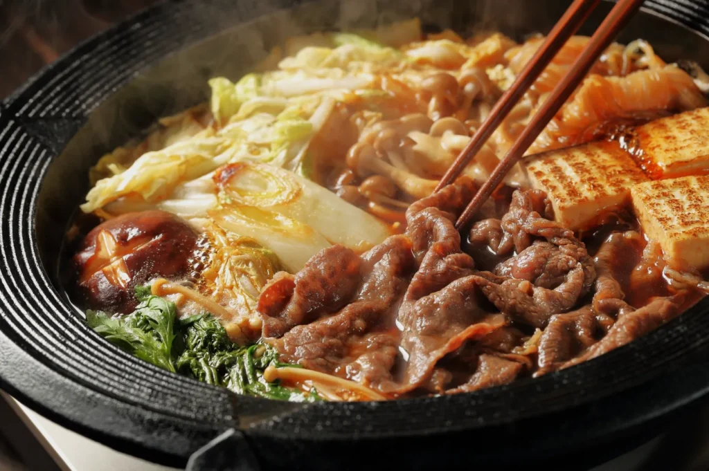 A steaming hot pot filled with thinly sliced beef, tofu, mushrooms, green vegetables, and cabbage, stirred with chopsticks, conveys warmth and comfort.