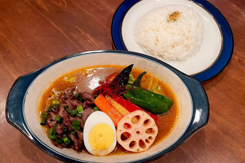 A vibrant bowl of Japanese curry with sliced beef, boiled egg, assorted vegetables, and lotus root. Served with a side of white rice on a wooden table.