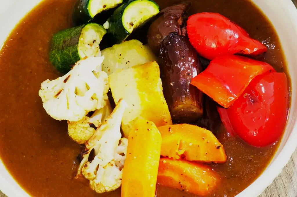A colorful bowl of curry features various vegetables, including red bell peppers, zucchini, cauliflower, and carrots, resting in rich brown sauce.