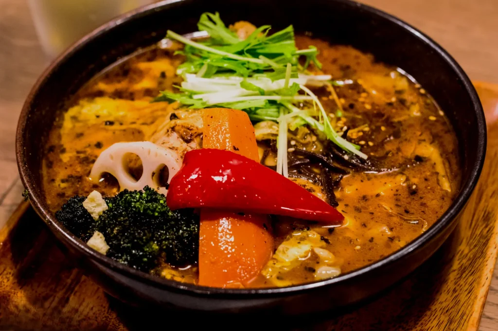 A vibrant bowl of Japanese soup curry features colorful vegetables like red pepper, carrot, broccoli, and lotus root, topped with fresh greens.