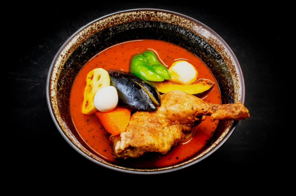 A rustic bowl of curry soup with a chicken drumstick, bright green pepper, boiled egg, lotus root, carrot, and eggplant, creating a colorful, appetizing display.