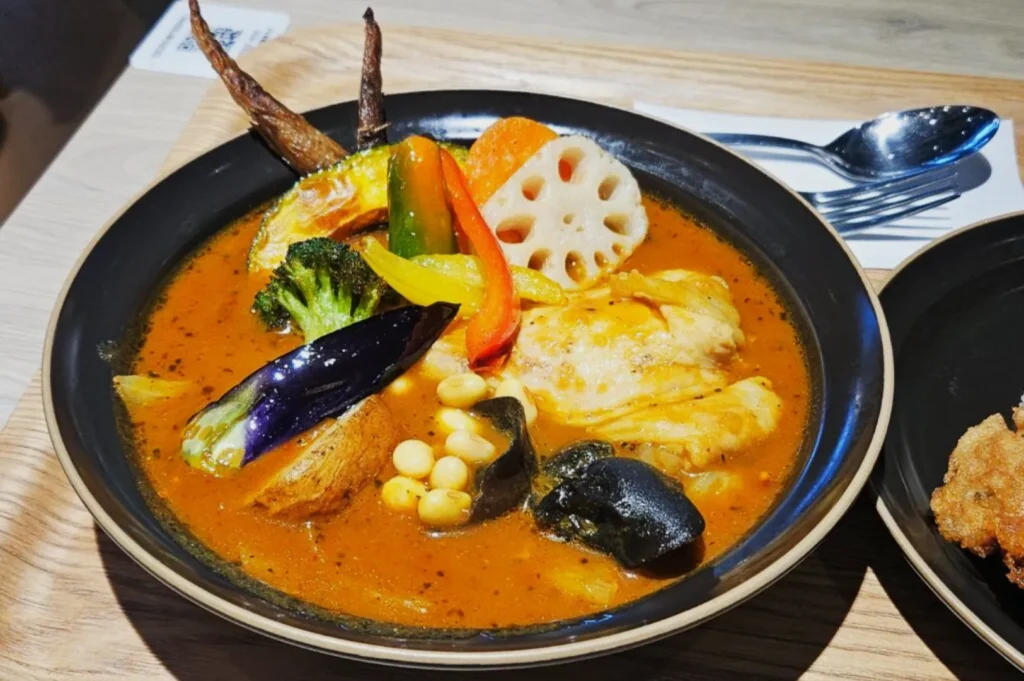 A vibrant bowl of Japanese curry soup with chicken, assorted vegetables, and beans in rich broth. Ingredients are artfully arranged for visual appeal.