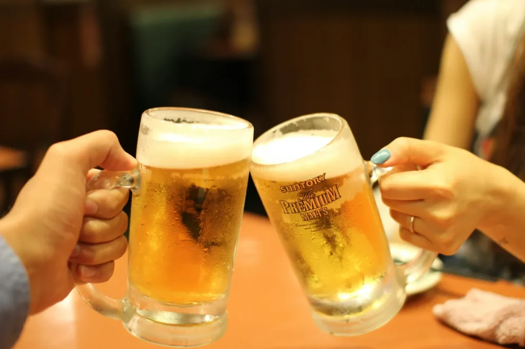 Two people clink large frosty beer mugs, filled with golden beer and topped with foam, creating a cheerful and celebratory atmosphere in a cozy setting.