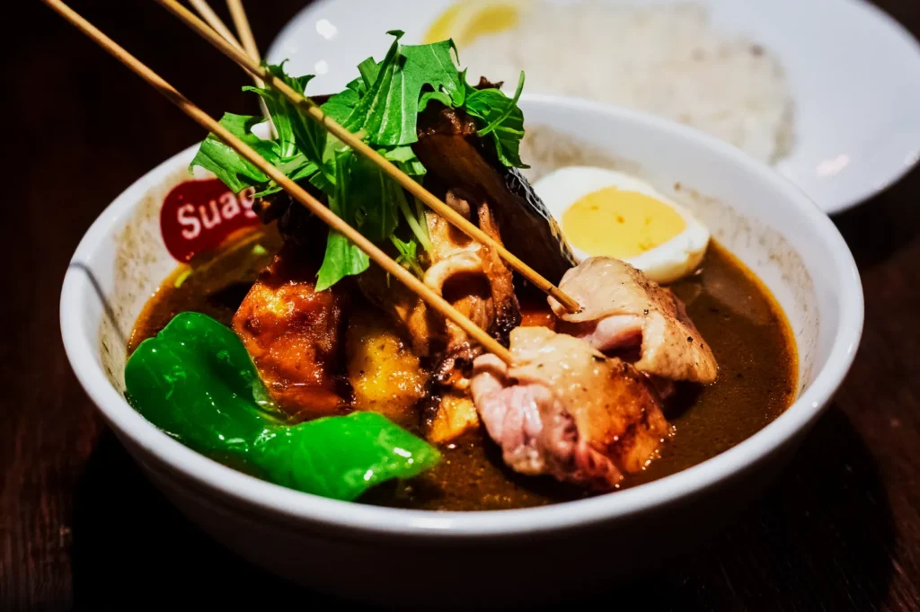 A bowl of flavorful Japanese soup curry with skewered meat, green pepper, a boiled egg, and leafy greens. A plate of rice is in the background.