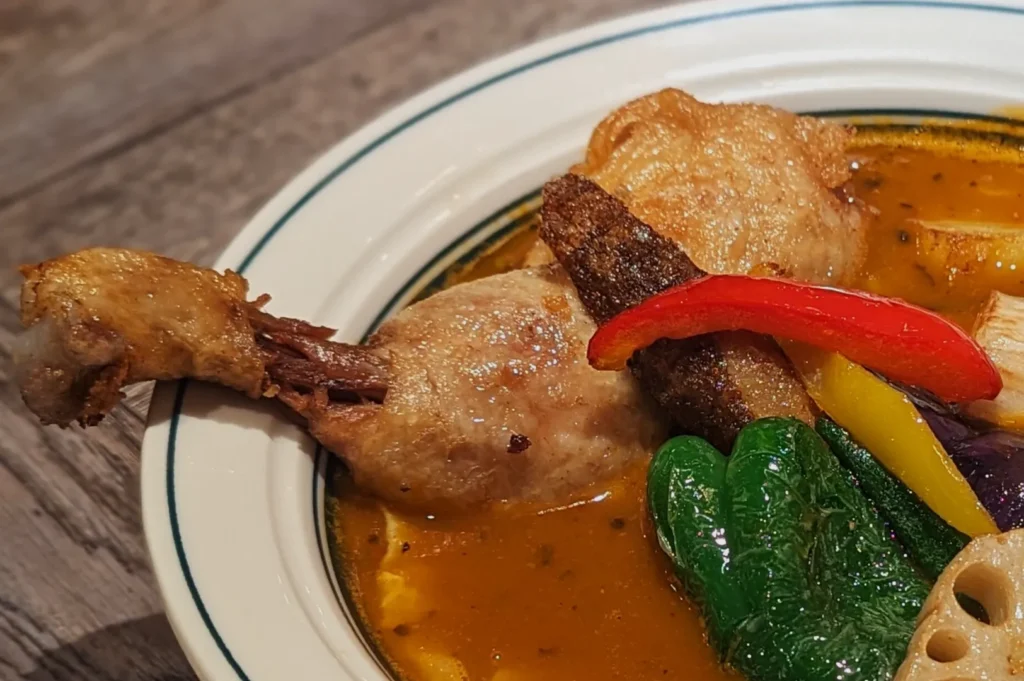 A bowl of colorful curry features a duck leg, vibrant red and yellow peppers, and green vegetables, creating a warm and inviting dish on a wooden table.