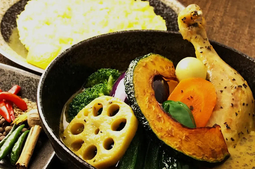 A vibrant bowl of chicken and vegetable curry includes colorful ingredients like pumpkin, broccoli, carrot, lotus root, and peppers, set beside yellow rice.