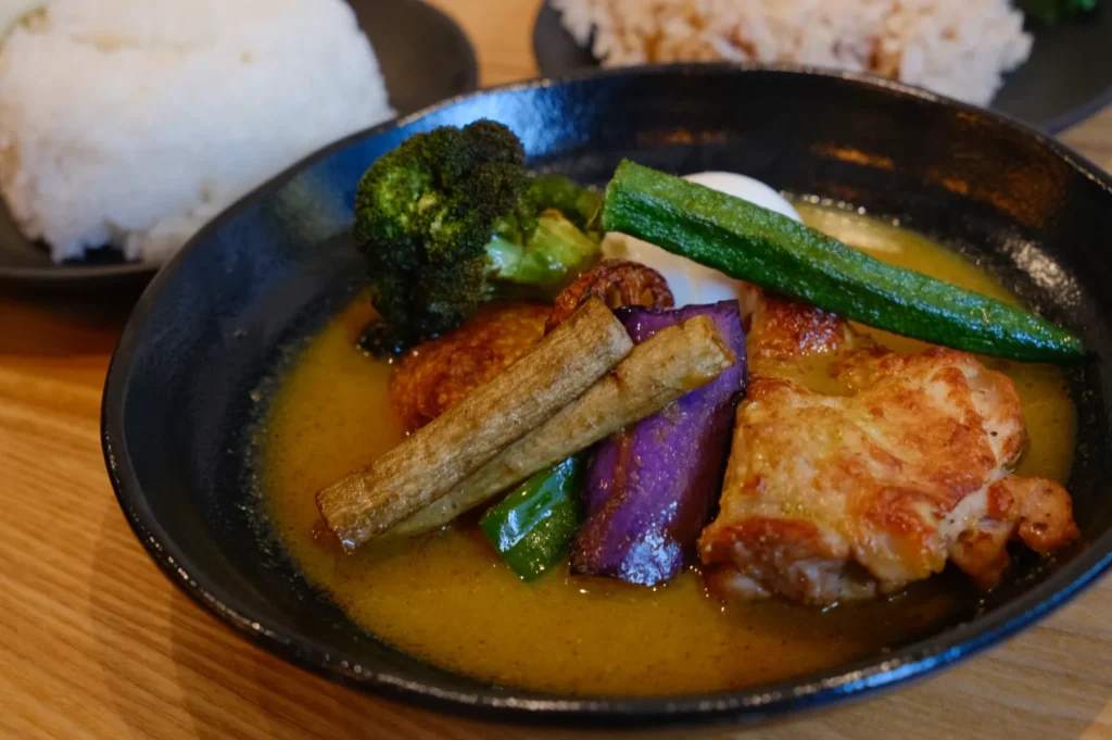 A bowl of curry featuring chicken, assorted vegetables including broccoli, eggplant, and okra, served with a side of rice.