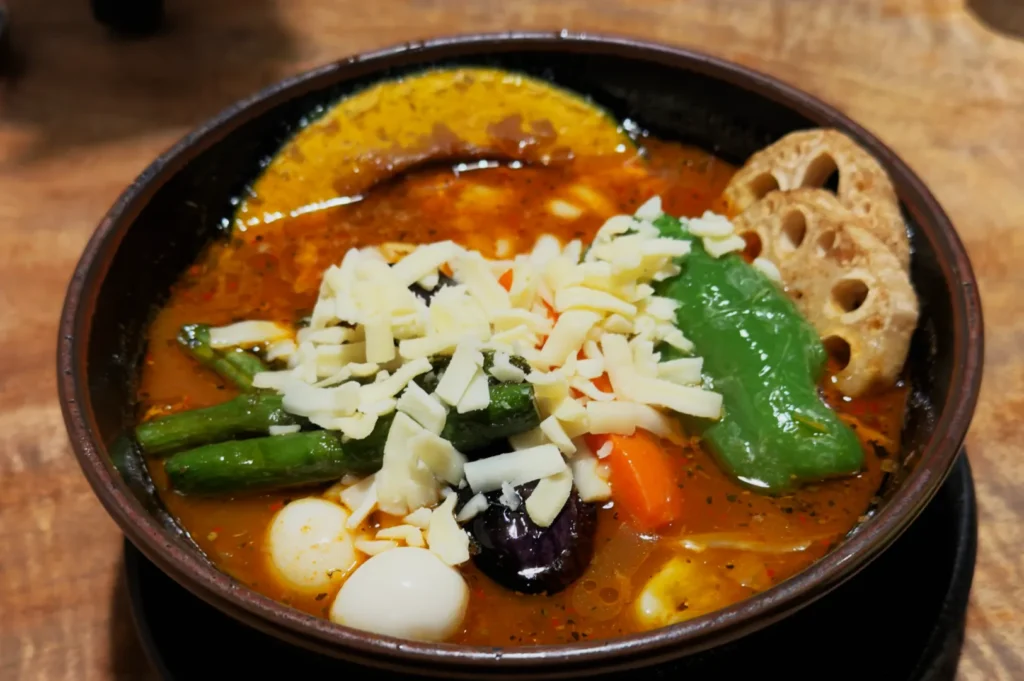 A bowl of vibrant vegetable stew with green beans, bell pepper, eggplant, quail eggs, and lotus root, topped with shredded cheese, on a wooden table.