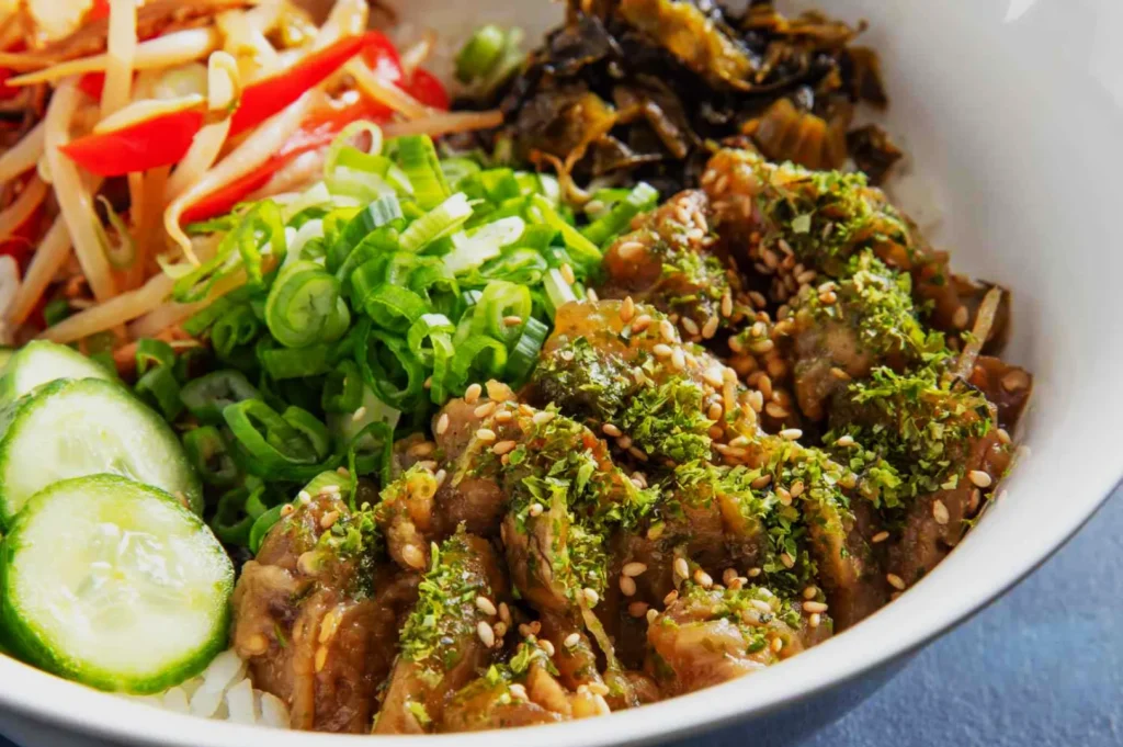A vibrant dish with sliced beef topped with green herbs and sesame seeds, scallions, pickled vegetables, sliced cucumbers, and peppers on rice.