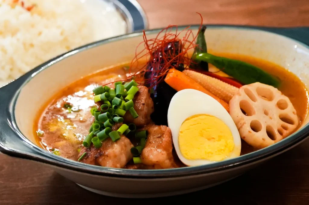 A vibrant bowl of Japanese curry soup featuring chicken pieces, a halved boiled egg, lotus root, carrot, and green chili, garnished with green onions.