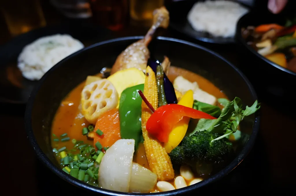 A vibrant bowl of curry with a chicken leg, assorted colorful vegetables, and lotus root in rich broth. Rice is in the background, creating a warm, appetizing scene.