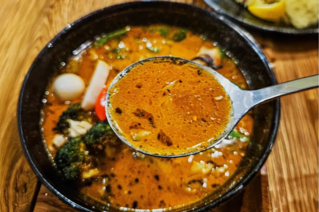 A close-up of a spoon lifting vibrant, orange-red broth from a bowl of spicy seafood soup, with visible vegetables and seafood chunks. Warm, savory feel.