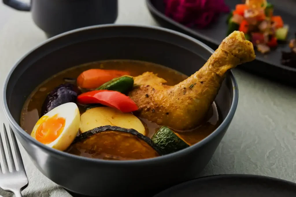 A bowl of soup curry with a chicken drumstick, boiled egg, carrot, bell peppers, potatoes, and zucchini in rich broth. A plate of vegetables is in the background.