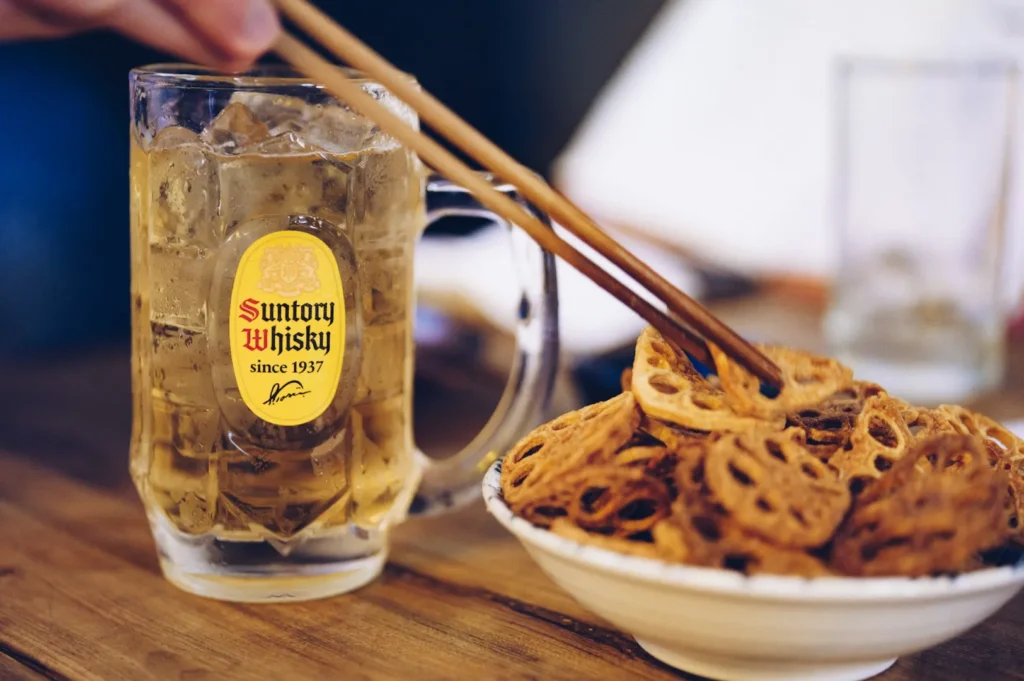 A mug of Suntory Whisky with ice sits on a wooden table next to a bowl of crispy lotus root chips, as chopsticks pick up a chip. Cozy and inviting.