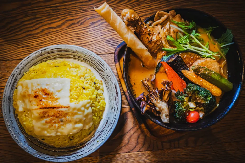 A bowl of yellow rice topped with cheese and spices, next to a platter of colorful curry with vegetables and crisp breadsticks, on a wooden table.