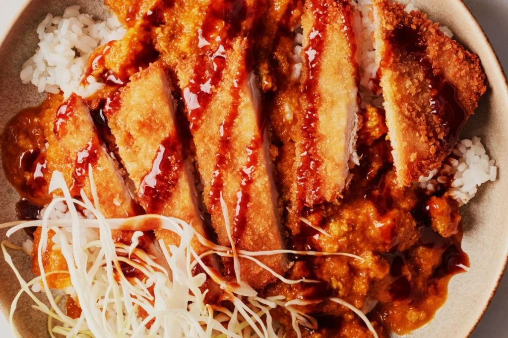 A plate of crispy, golden-brown breaded chicken slices drizzled with dark sauce, served on white rice and topped with shredded cabbage and curry sauce.