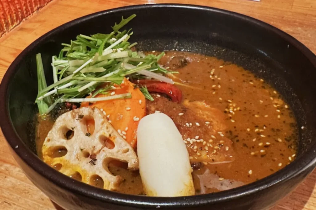 A black bowl of hearty soup with vibrant ingredients on a wooden table. Topped with lotus root, carrot, greens, and potato in a rich broth.