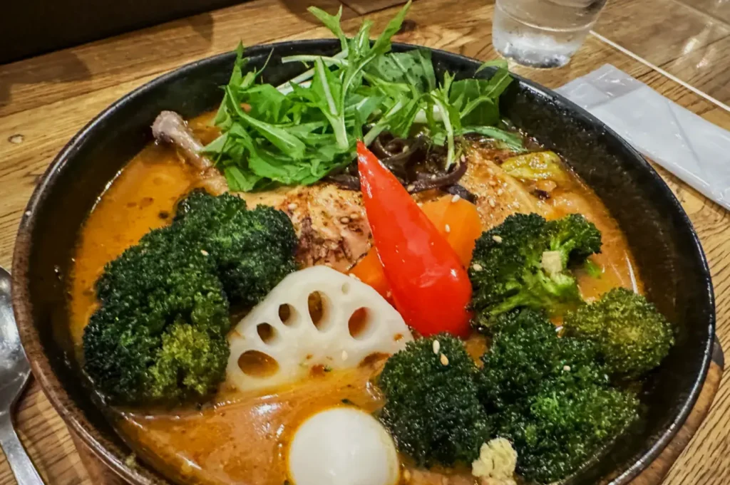 A vibrant bowl of ramen with rich, orange broth, topped with broccoli, lotus root, leafy greens, a bright red pepper, and a boiled egg on a wooden table.