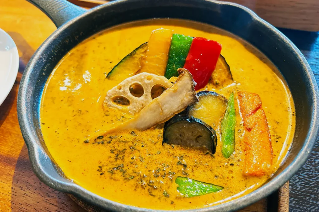 A bowl of vibrant yellow curry soup filled with assorted colorful vegetables including red, green, and yellow peppers, lotus root, and eggplant, creating a warm and appetizing appearance.