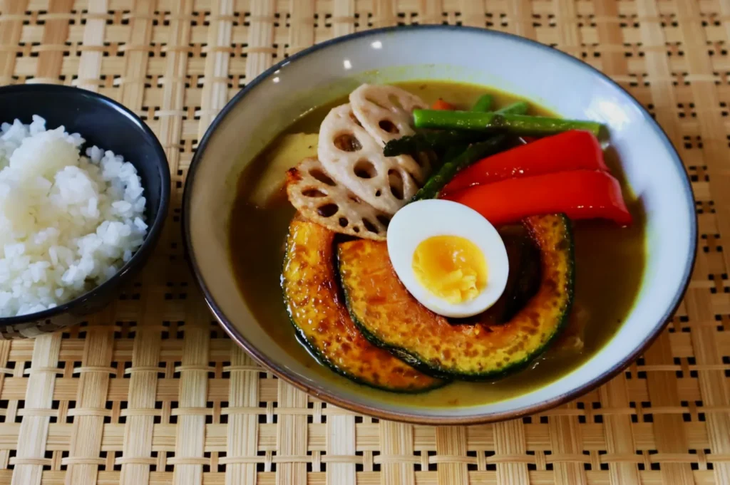 A bowl of soup curry with vibrant slices of pumpkin, lotus root, red pepper, asparagus, and a halved boiled egg, beside a bowl of white rice.