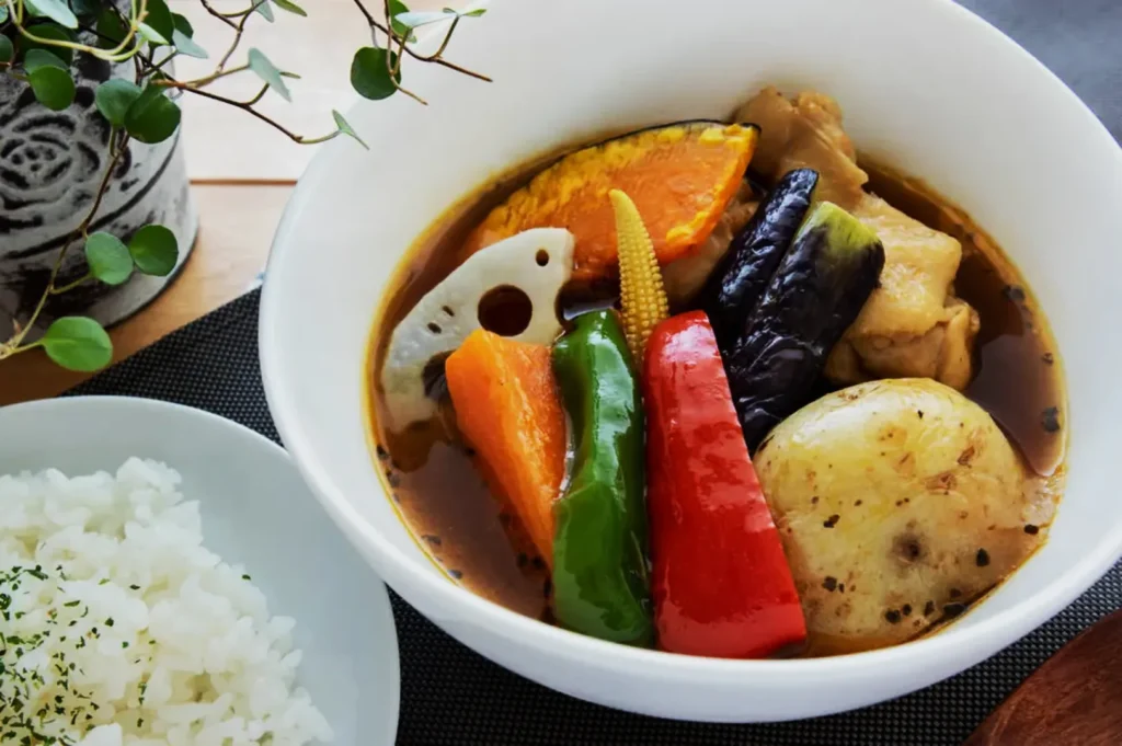 A bowl of hearty soup with colorful vegetables including pumpkin, baby corn, lotus root, and bell peppers, next to a plate of white rice, garnished with herbs.