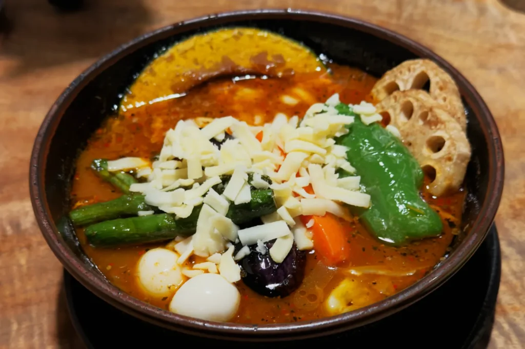 A bowl of vibrant, spicy curry soup topped with sliced cheese, quail eggs, green peppers, asparagus, lotus root, and vivid vegetables on a wooden table.