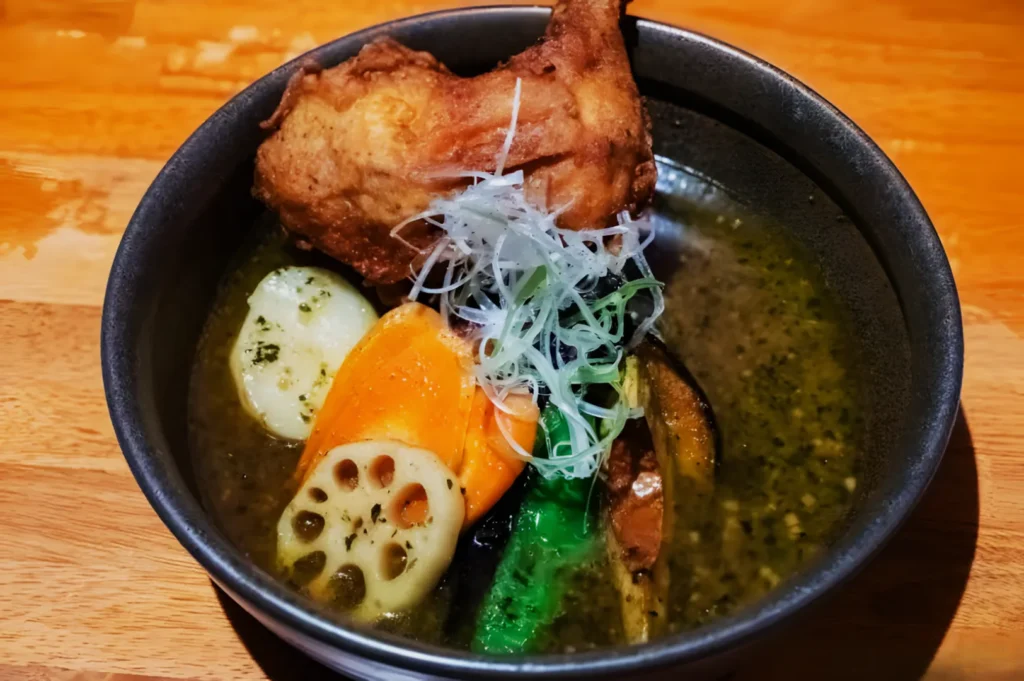 A bowl of Japanese soup curry with a piece of fried chicken, sliced carrots, lotus root, green beans, and shredded white onions on top.