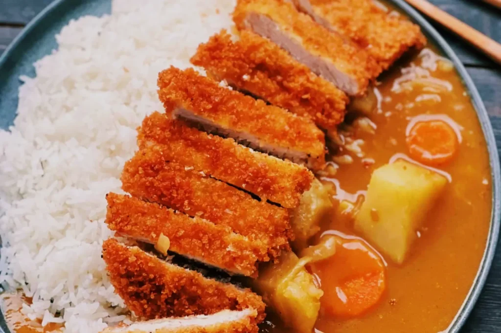 Plate of Japanese katsu curry with crispy breaded cutlets, white rice, and rich curry sauce featuring potato and carrot chunks, conveying comfort and warmth.