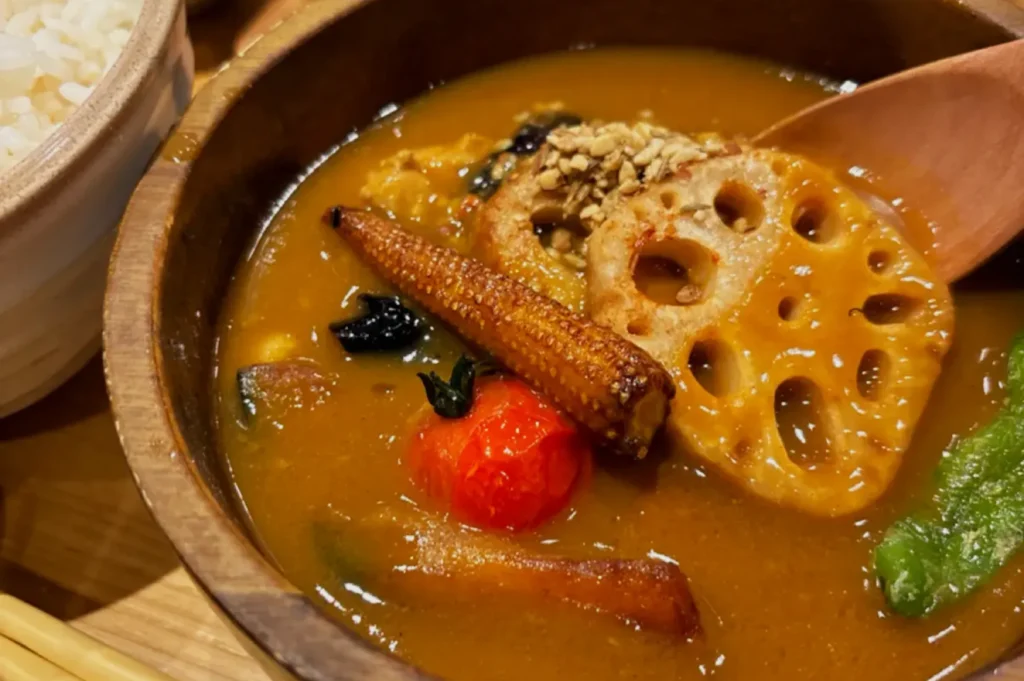 A vibrant bowl of curry soup with lotus root, baby corn, cherry tomato, greens, and sesame seeds. A wooden spoon rests inside. Warm, appetizing scene.