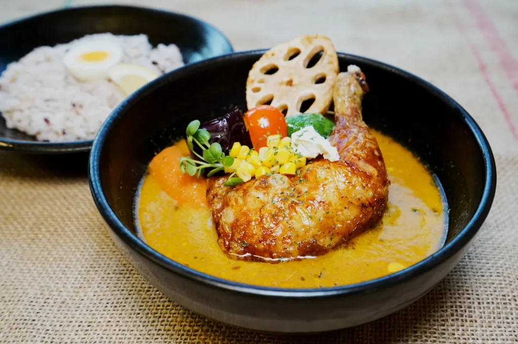 A bowl of chicken curry topped with vegetables like corn, lotus root, and greens on a creamy sauce. A side dish of rice and boiled eggs is visible.