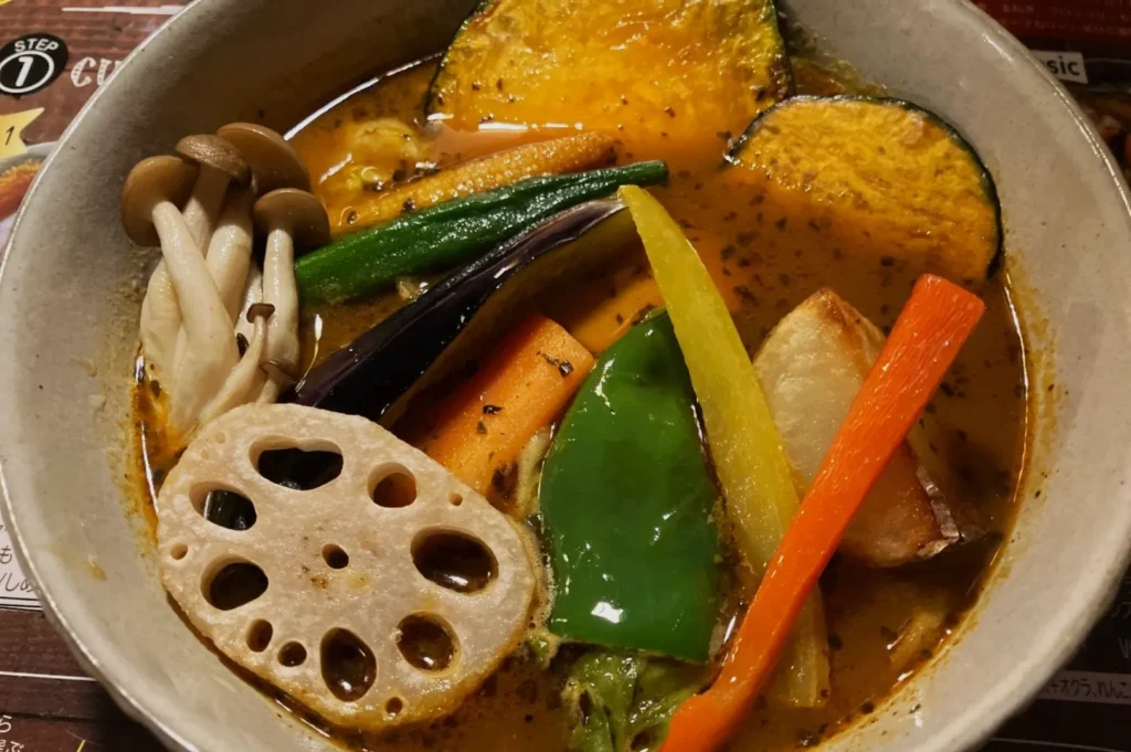 A bowl of colorful vegetable curry with lotus root, mushrooms, bell pepper, squash, and carrots. The dish has a warm, hearty appearance.
