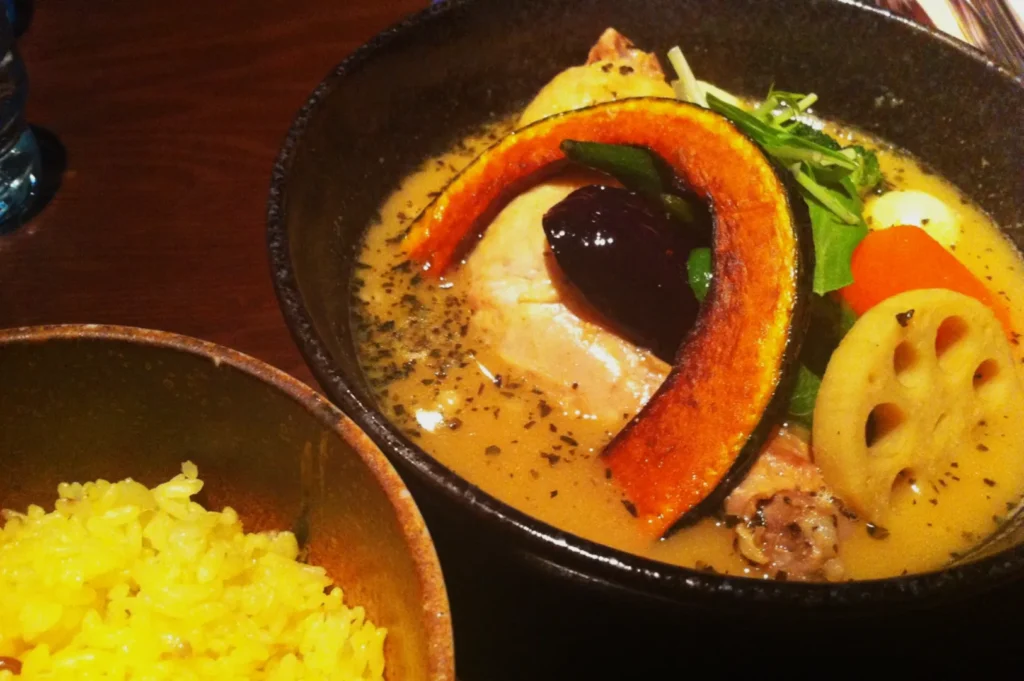A bowl of creamy soup curry with chicken, vegetables, and herbs sits beside a bowl of yellow rice on a wooden table, evoking a cozy dining atmosphere.