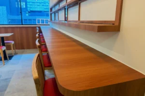 A wooden countertop with a shelf above in a cafe, featuring red chairs and a large window with a city view.