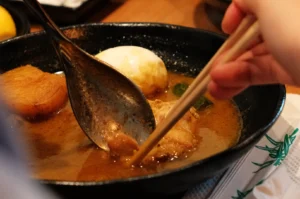 A hand uses chopsticks to hold a piece of meat in a rich, brown broth in a black bowl. A spoon and boiled egg are also visible, creating a cozy, appetizing scene.