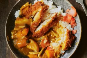 A bowl of Japanese curry with rice, topped with slices of crispy fried chicken. Carrots and potatoes are visible, along with red pickled ginger.