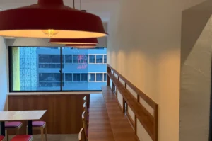 Modern dining area with wooden tables and chairs, red pendant lights, and a large window overlooking an urban cityscape. Cozy and inviting atmosphere.