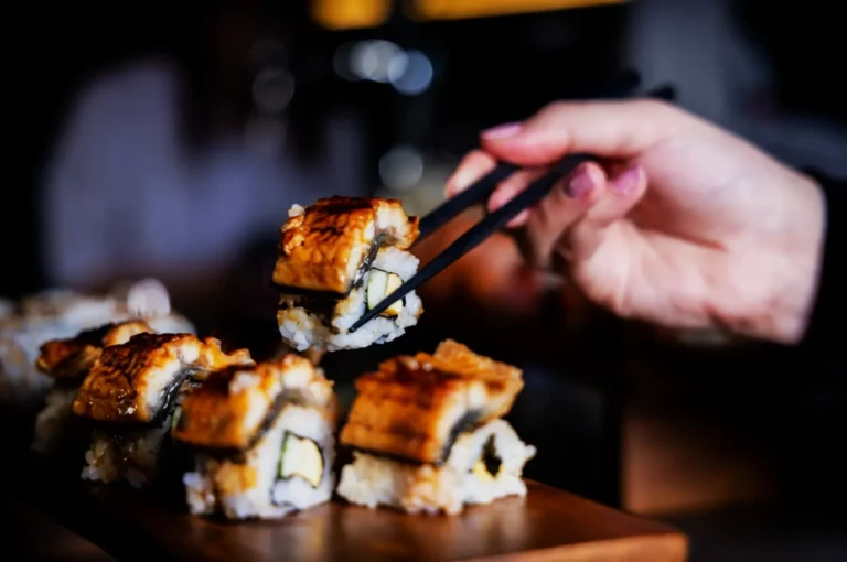 A hand uses chopsticks to pick up a sushi roll topped with eel from a wooden board. The sushi is glossy, highlighting its freshness and deliciousness.