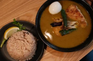 A bowl of curry featuring a piece of chicken, a boiled egg, and green vegetables, alongside a serving of rice and a lemon wedge on a plate.